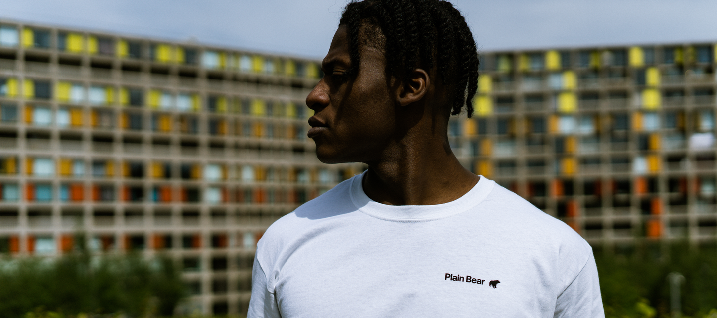 white tee with black plain bear text logo. modelled by a black male in front of a block of colorful flats